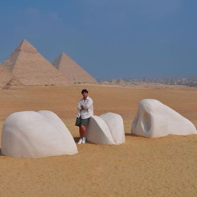 Jwan Yosef is sitting on the lips of the structure with Pyramids in the background.